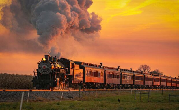 A View of an Antique Steam Passenger Train Approaching at Sunrise With a Full Head of Steam and Smoke Traveling Thru Farmlands
